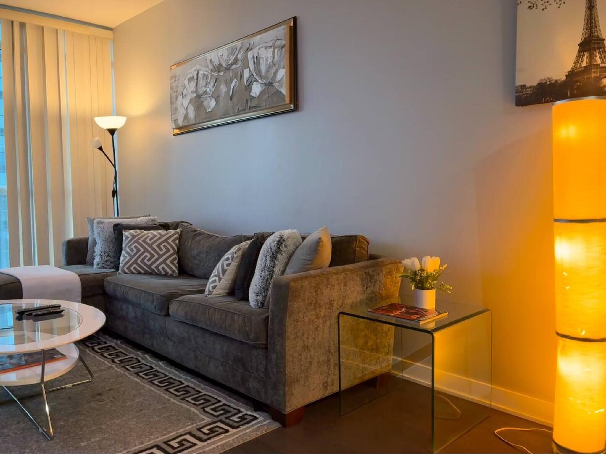 Una sala de estar moderna con un sofá gris, una mesa de centro de vidrio, un tapete estampado y un cuadro de flores.