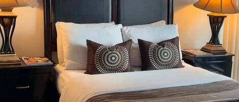 Una cama bien tendida con un cabecero oscuro, dos mesitas de noche con lámparas y almohadones decorativos.