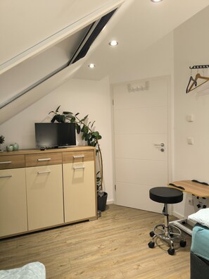 A compact room with a sloped ceiling, a wooden desk with a chair, a television on a cabinet, and a door with a coat rack.