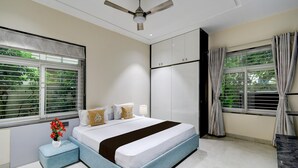 A bedroom with a bed, a ceiling fan, a window with blinds, and a nightstand with a vase of flowers. - Villa Verizon by Dancenter (Hyderabad)