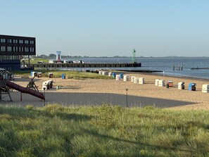 A beach with beach huts, a playground, a pier, and a building in the background. - Skipper's Deck: Modern, near harbor & dike, incl. parking, 2 workstations, wifi (Bremerhaven)