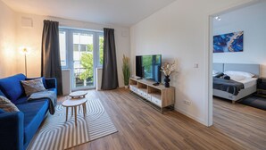 En moderne stue med blå sofa, fjernsyn på træfod, seng og stort vindue med gardiner.