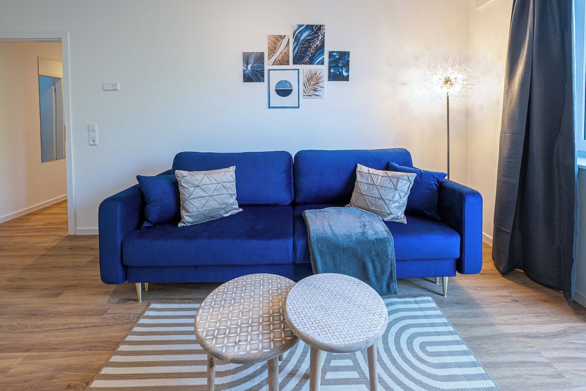 Een moderne woonkamer met een blauwe bank, twee bijzettafeltjes met dessin en een decoratief wandobject.