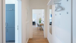 En moderne gang med trægulv, hvide vægge og en bænk. Der er en dør, der fører til et køkken med et blå køleskab.