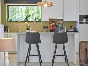Una cucina moderna con mobili bianchi, un'ampia finestra con piante e un'opera d'arte a tema frutta, più due sgabelli da bar.