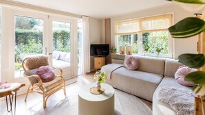 Ein geräumiges Wohnzimmer mit Sofa, Sessel, Fernseher und einem großen Fenster mit Blick auf Grünflächen.