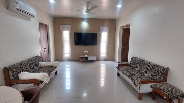A spacious living room with two patterned sofas, a flat-screen TV mounted on the wall, and a ceiling fan.