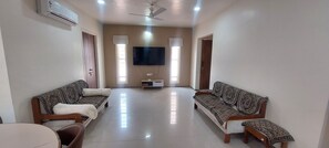 A spacious living room with two patterned sofas, a flat-screen TV mounted on the wall, and a ceiling fan. - Family home near Airport & convention center, withFree Wi-Fi & aminities. (Surat)