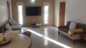 A living room with a sofa, a TV, a dining table with a bottle, and a window with blinds.