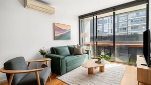Un soggiorno moderno con un divano verde, un tavolino in legno e una poltrona grigia. Si apre su un'ampia finestra con vista su un edificio e un balcone.