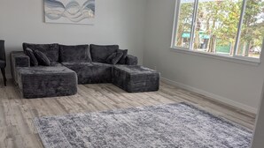 A modern living room with a sectional sofa, a large rug, and a view of greenery outside. - Newly built 3-bedroom house in beautiful Hilo perfect for a relaxing stay. (Hilo)