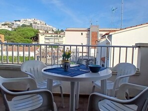 Ein Balkon mit rundem Tisch, zwei Stühlen, einem Blumentopf und Blick auf eine Hügelstadt.