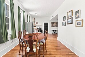 A dining area with a wooden table and chairs, a kitchen in the background, and artwork on the walls.