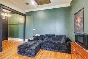 Uma sala de estar com sofá seccional, piso de madeira, parede verde escuro e um quadro.