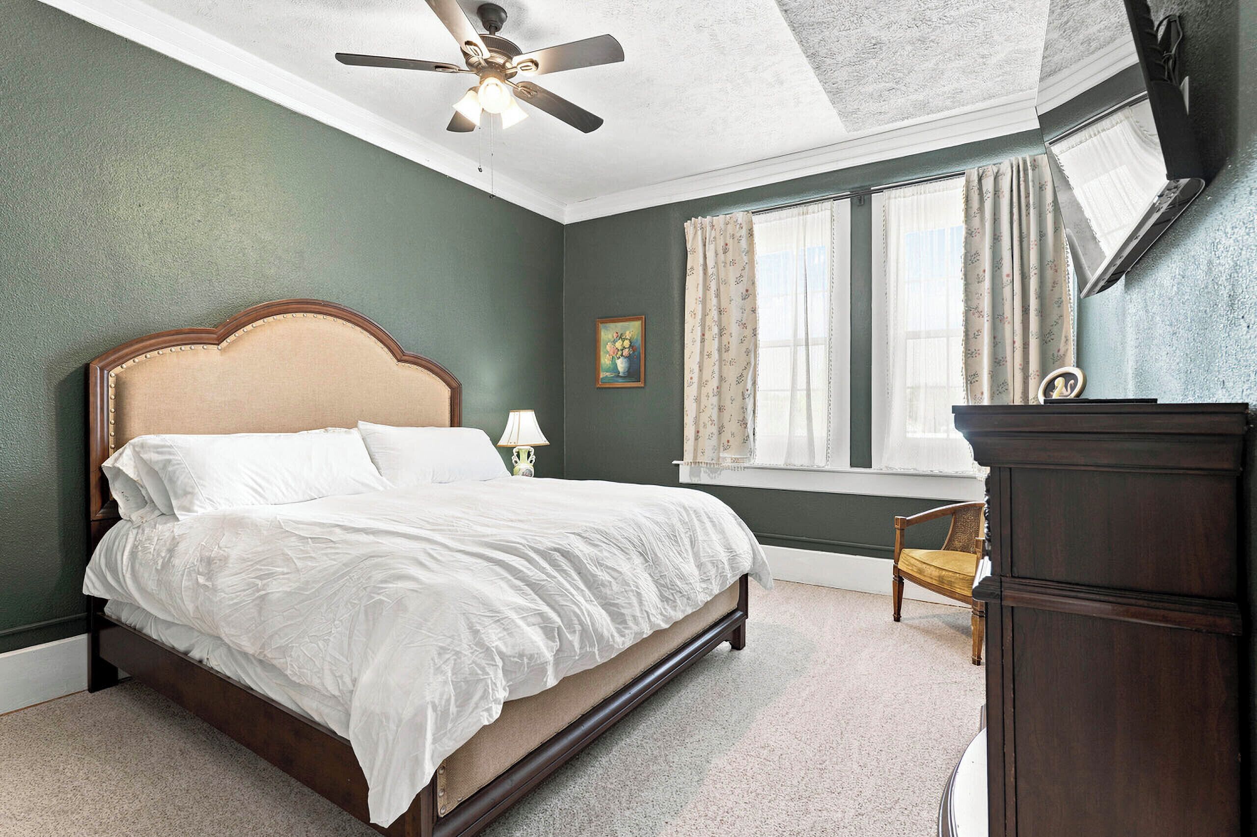 Une chambre à coucher comprenant un lit, une table de chevet, une commode, une chaise, une fenêtre avec des rideaux, un ventilateur de plafond et un tableau accroché au mur.