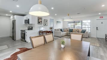 A modern kitchen with stainless steel appliances, a dining area with a wooden table and chairs, and a living room with a sofa and a television.