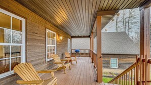 En träveranda med Adirondack-stolar, ett takförsett område med ett räcke och en utsikt över ett hus i bakgrunden.