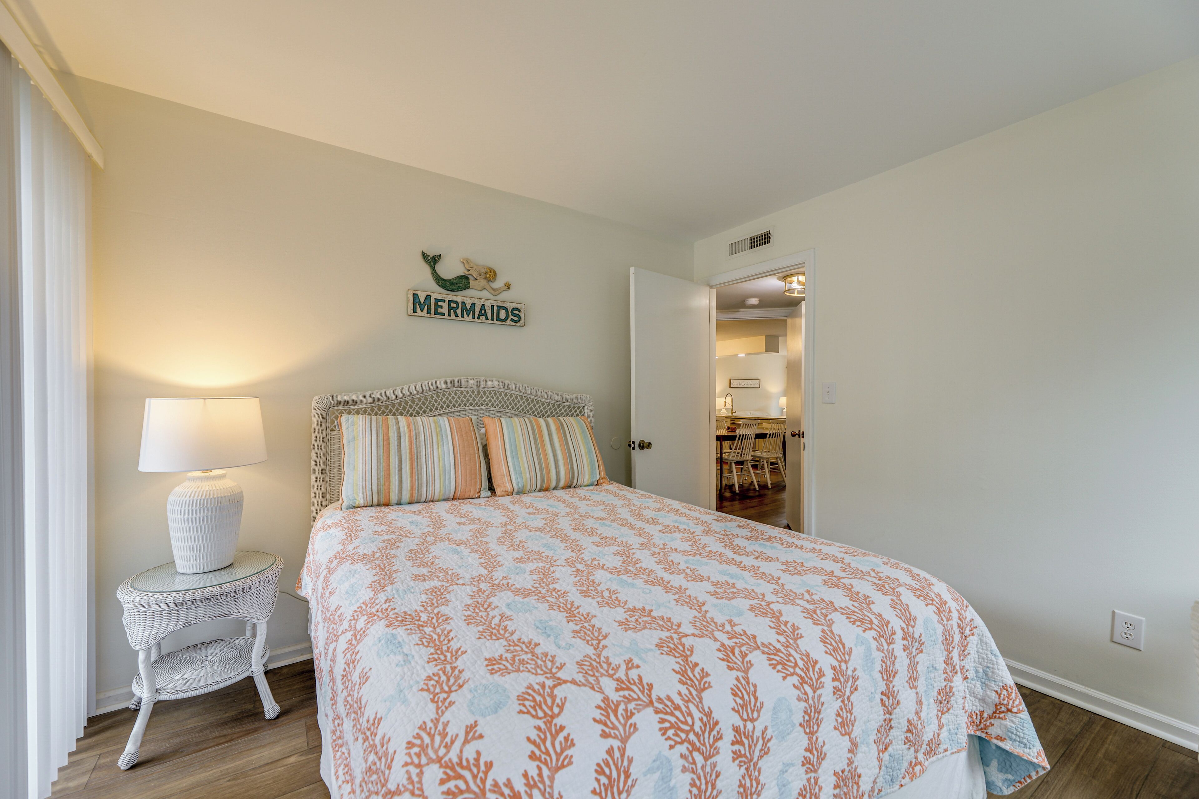A bedroom with a bed, a nightstand with a lamp, and a sign on the wall that says "MERMAIDS".