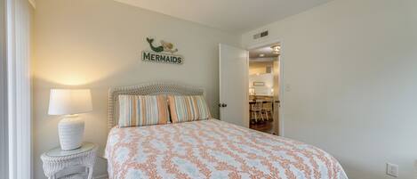 A bedroom with a bed, a nightstand with a lamp, and a sign on the wall that says "MERMAIDS".