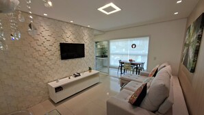 Ein modernes Wohnzimmer mit weiß gefliestem Boden, einem weißen Sofa mit bunten Kissen, einer weißen Unterhaltungsanlage und einem Flachbildfernseher an einer strukturierten Wand.