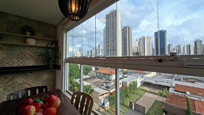 Ein Zimmer mit großem Fenster und Blick auf eine Stadtlandschaft, einem Esstisch mit Äpfeln und einer hängenden Leuchte.