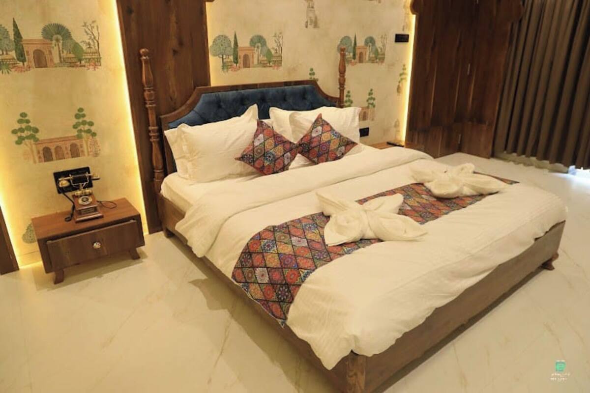 A double bed with a patterned bedspread and pillows, a wooden nightstand with a telephone, and a wall with a mural.