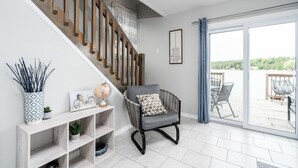 A modern living room with a staircase, a grey armchair, a console table with decorative items, and a view of the outdoors through large glass doors. - Scenic Waterfront Cottage Retreat - Firepit & BBQ (Hanover)