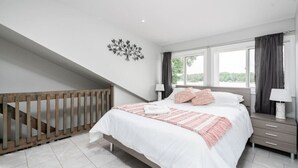 A bedroom with a bed, a nightstand, a lamp, a window with curtains, a staircase, and a decorative wall piece. - Scenic Waterfront Cottage Retreat - Firepit & BBQ (Hanover)