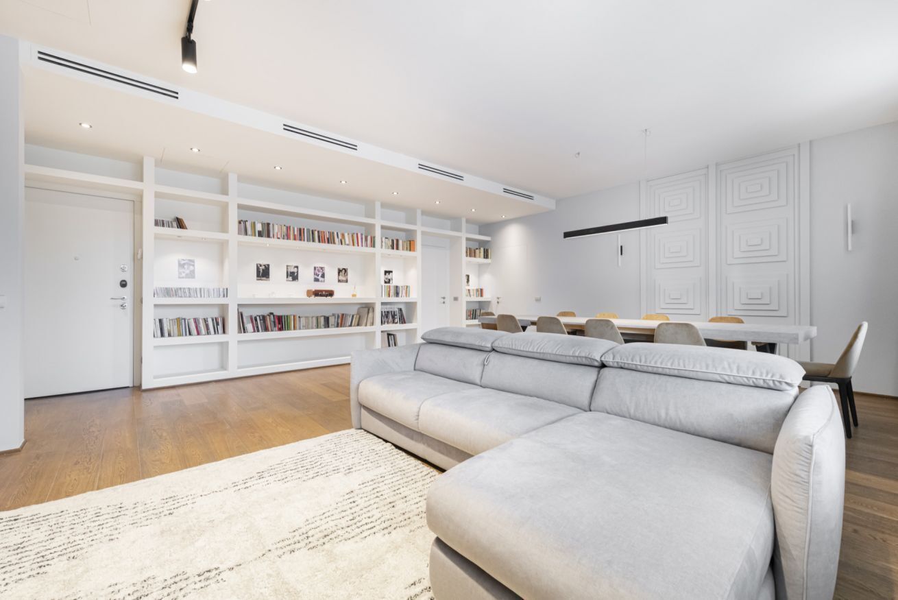 Een moderne woonkamer met een grijze hoekbank, witte inbouwboekenplanken en een eethoek met een witte tafel en stoelen.