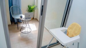 Une pièce avec une porte-fenêtre coulissante, une chaise blanche, une petite table avec un ordinateur portable, une plante en pot et un coussin jaune sur un banc blanc.