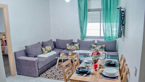 Un salon avec un canapé gris, une table à manger en bois pour quatre personnes et une fenêtre ornée de rideaux verts.