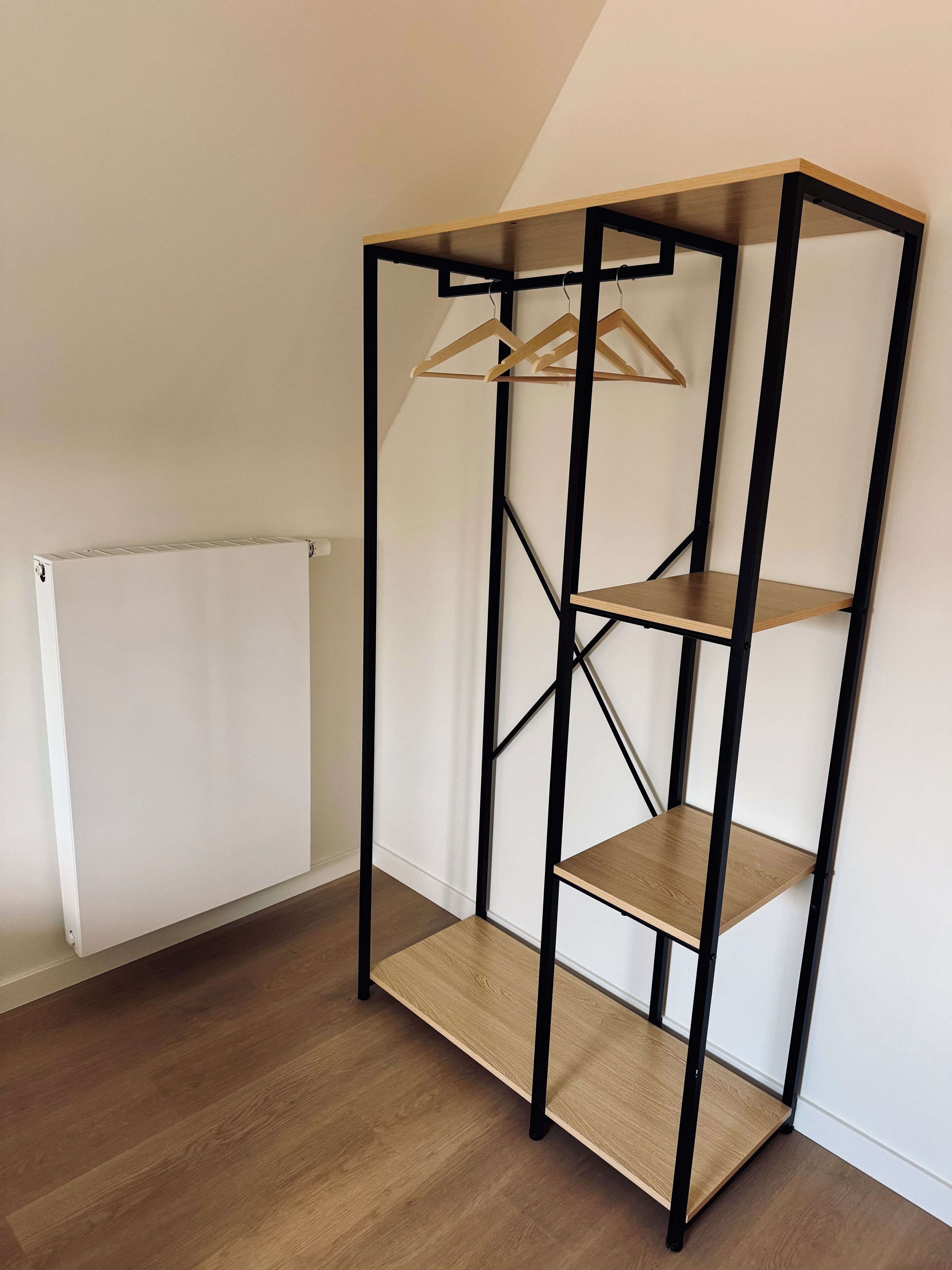 A corner room with a wooden floor, a radiator, and a black metal shelving unit with wooden shelves.