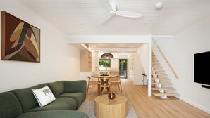 Ein modernes Wohnzimmer mit einem grünen Sofa, einem runden Couchtisch, einem Essbereich mit Tisch und Stühlen, einer Treppe und einem an der Wand befestigten Fernseher.