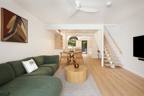 A modern living room with a green sofa, a round wooden coffee table, a dining area with a table and chairs, a staircase, and a TV mounted on the wall. - Ray St Retreat - Luxury Stay near Sunshine Beach (Sunshine Beach)