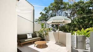 Eine Terrasse mit einer Couch, einem runden Tisch und einem Sonnenschirm, umgeben von Grünpflanzen und einer weißen Wand.