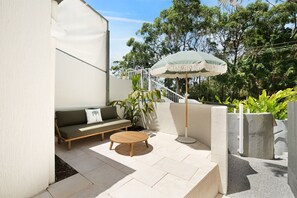 A patio with a sofa, round table, and umbrella, surrounded by greenery and a white wall. - Ray St Retreat - Luxury Stay near Sunshine Beach (Sunshine Beach)