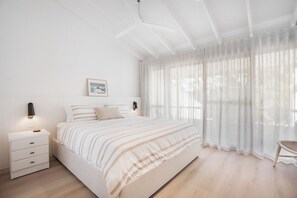 A bedroom with a large bed, a bedside table with drawers, a wall-mounted light, and sheer curtains. - Ray St Retreat - Luxury Stay near Sunshine Beach (Sunshine Beach)