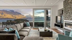 Un soggiorno moderno con un divano ampio, una televisione a schermo piatto e un camino. C'è un balcone con vista sulle montagne e su un lago.