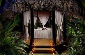 A thatched-roof gazebo with a white table set for a meal, surrounded by tropical plants.