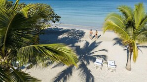 A beach with palm trees, a clear blue sea, and two people walking. There are lounge chairs and a small pool area. - Couples Negril - Adults Only (Negril)