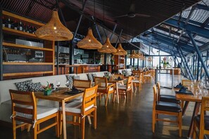 Un restaurante con mobiliario de madera, lámparas de mimbre colgantes y un techo alto con vigas de metal.