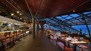 Un restaurant doté d’un long comptoir et de chaises en bois, d’un plafond haut avec une charpente en bois, et offrant une vue sur la mer.
