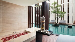 Une salle de bain moderne avec une baignoire, un plancher en bois, une petite table avec des tasses et une cage à oiseaux, et une vue sur une piscine et un bâtiment en arrière-plan.