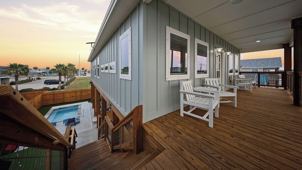 A covered deck with wooden flooring, white outdoor furniture, and a view of a swimming pool and distant buildings. - Private Pool + Putting Green + Golf Cart Zone + Walk to Beach | Yahwaves (Port Aransas)