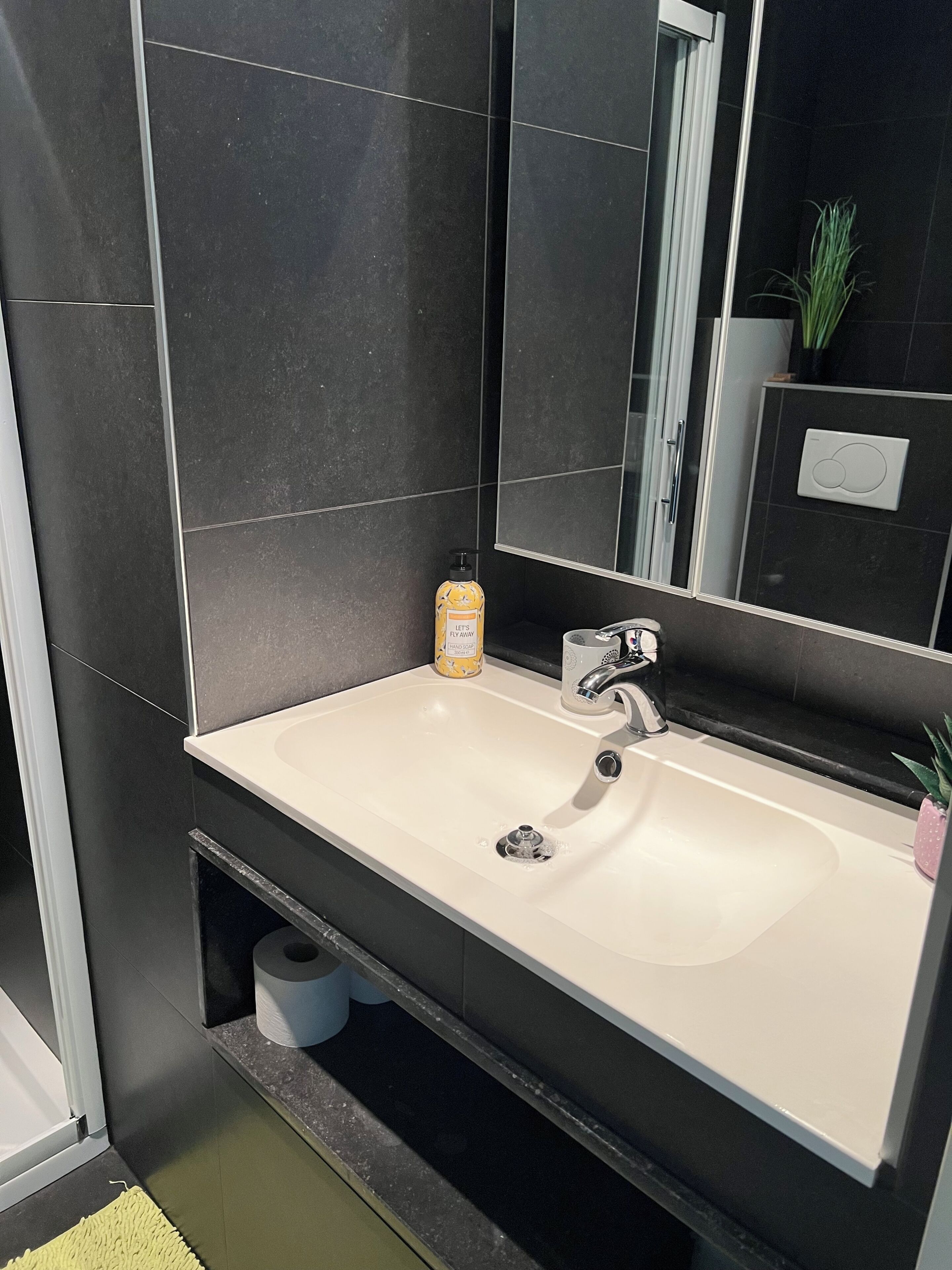 Un baño moderno con un lavamanos blanco, baldosas oscuras, un espejo y un estante pequeño con artículos de aseo.