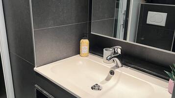 Un baño moderno con un lavamanos blanco, baldosas oscuras, un espejo y un estante pequeño con artículos de aseo.