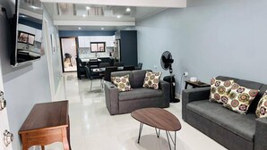 Un salón moderno con un sofá gris, una mesa de centro de madera y cojines estampados. En el fondo, se observa un comedor con una mesa y sillas negras.