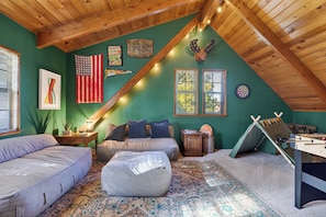 Ein gemütliches Schlafzimmer im Dachgeschoss mit einem Hochbett, einer Futonliege, einem Futonsessel, einem kleinen Tisch, einer Lampe, einer Wanduhr, einem Wandbild und einem Kicker.