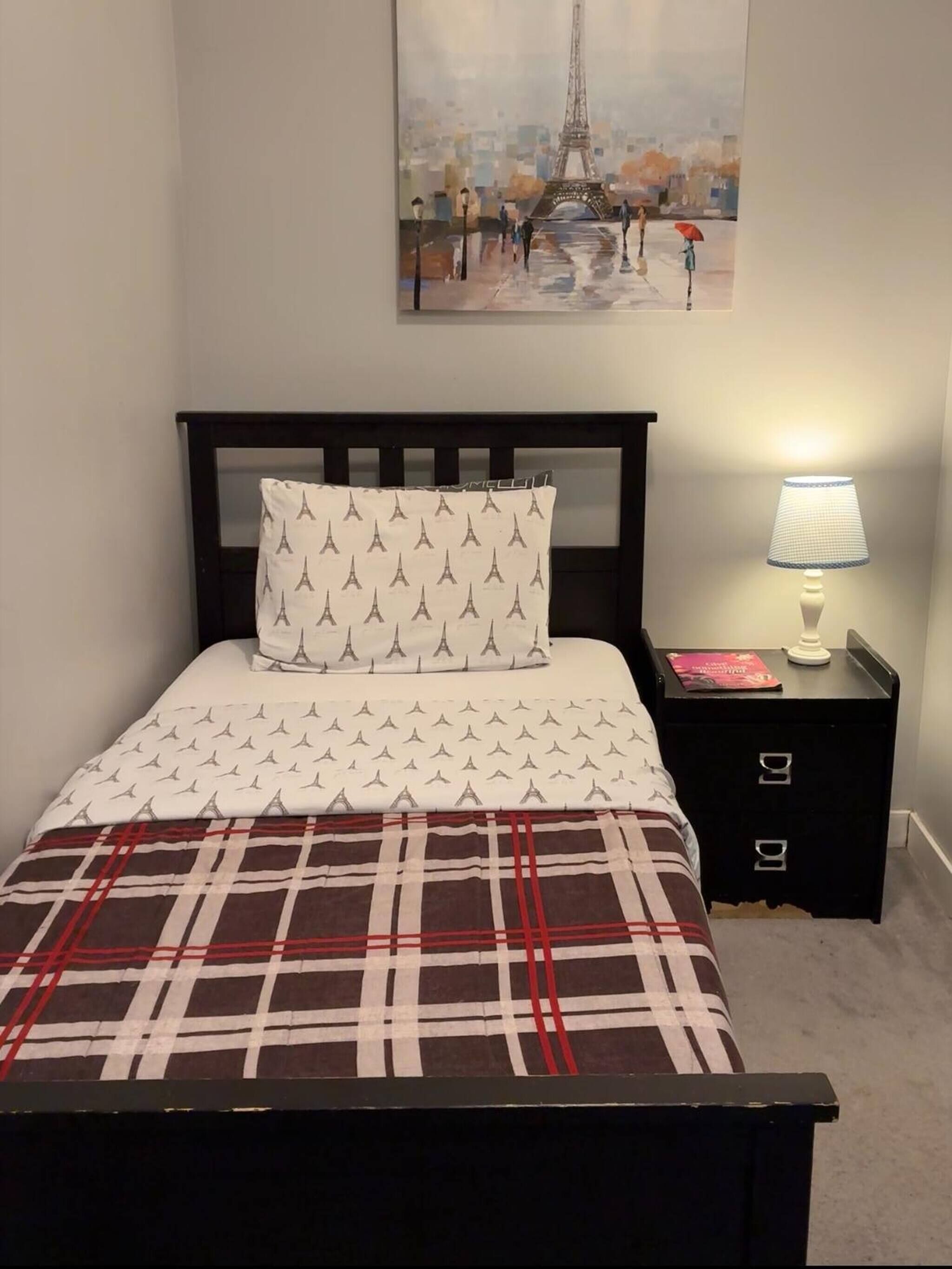 Een slaapkamer met een bed, nachtkastje, lamp en een schilderij van de Eiffeltoren.