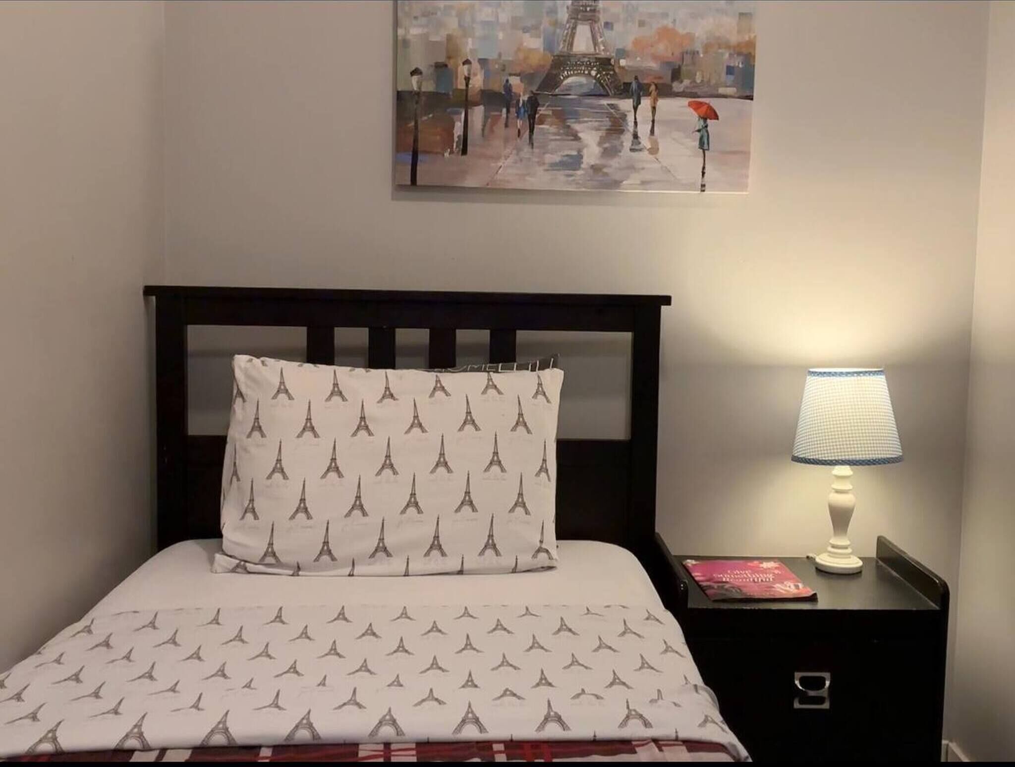Een hotelkamer met een bed met een wit en grijs patroon van de Eiffeltoren, een donker houten hoofdbord, een nachtkastje met een lamp en een schilderij van de Eiffeltoren aan de muur.